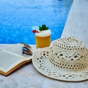 beige straw hat book sunglasses and drink beside pool
