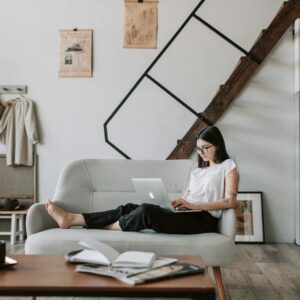 Positive female with tattooed arms wearing casual clothes and eyeglasses reading netbook while sitting barefoot on comfortable sofa behind table with books and magazines in modern apartment with minimalist interior in eco style during free time