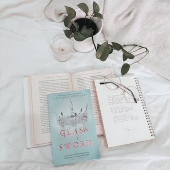 A cozy reading nook featuring the book 'Glass Sword', glasses, and a notepad on a bed.