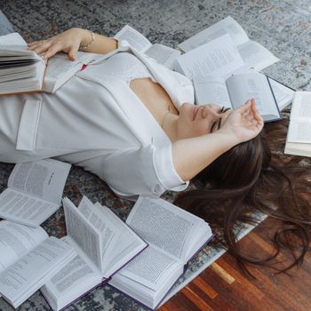 A woman in a white outfit lying on a rug surrounded by open books, suggesting relaxation and learning.