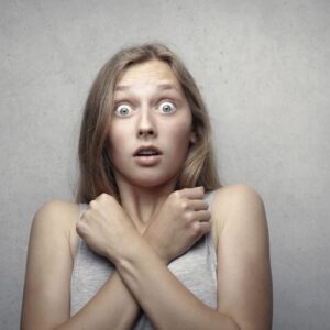 Portrait of a surprised woman with wide eyes and crossed arms against a gray background. syndrome de l'imposteur chez l'auteur