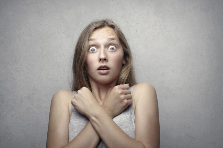 Portrait of a surprised woman with wide eyes and crossed arms against a gray background. syndrome de l'imposteur chez l'auteur
