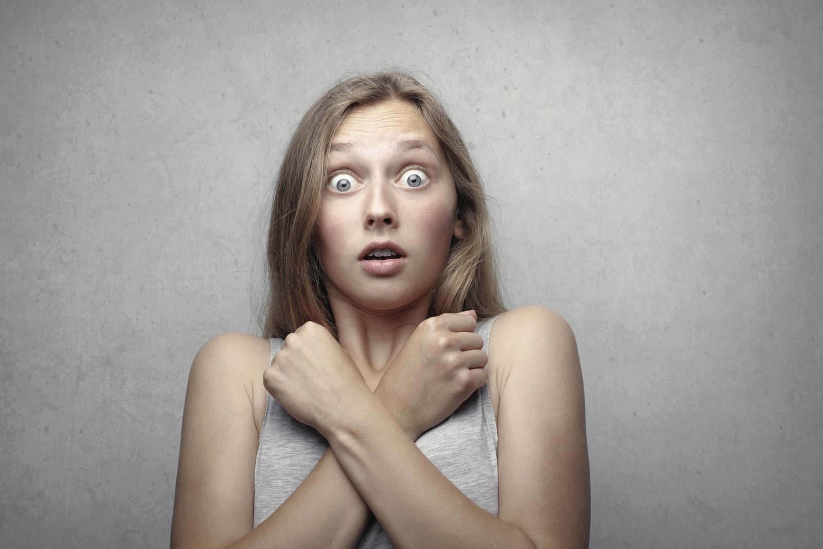 Portrait of a surprised woman with wide eyes and crossed arms against a gray background. syndrome de l'imposteur chez l'auteur