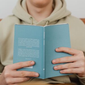 Close-up of hands holding a hardcover book, wearing a beige hoodie. Ideal for reading-related content.